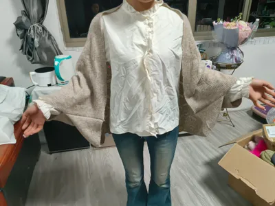 Frau trägt übergroßen beigen Strickjacke über cremefarbenem wattiertem Rüschenkragenhemd mit blauen Schlaghosen in Innenraum