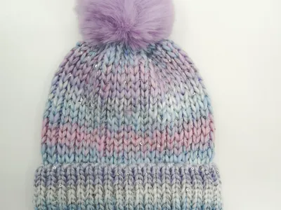 Grob gestrickte Beanie in Pastell-Ombre aus Lila, Blau und Rosa mit silbernen Pailletten und lavendelfarbenem Kunstfell-Bommel auf weißem Hintergrund