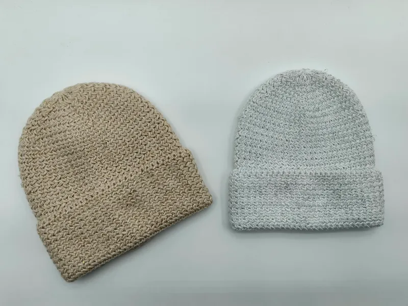 Zwei grob gestrickte Beanies nebeneinander: eine beige ohne Krempe und eine hellgraue mit umgeschlagenem Rand, auf weißem Hintergrund