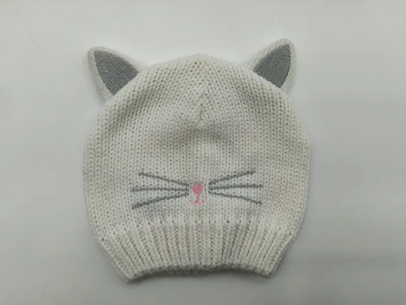 Weiße grob gestrickte Baby-Beanie mit silber glitzernden Katzenohren, aufgestickten Schnurrhaaren und rosa Nase auf grauem Hintergrund