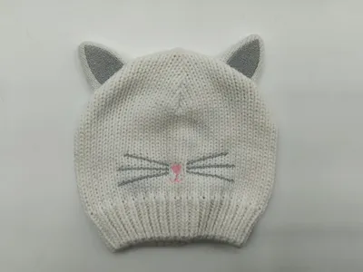 Weiße grob gestrickte Baby-Beanie mit silber glitzernden Katzenohren, aufgestickten Schnurrhaaren und rosa Nase auf grauem Hintergrund