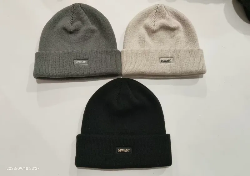 Drei Rippenstrickmützen mit gefaltetem Bund in Anthrazit, Cremeweiß und Schwarz auf weißer Oberfläche mit Logo-Patch