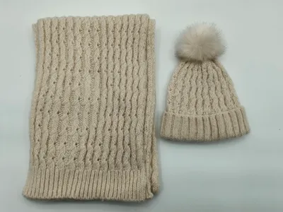 Cremefarbenes Winter-Set bestehend aus Zopfmuster-Beanie mit Kunstfell-Pompon und passendem Rippenstrickschal, flach ausgelegt