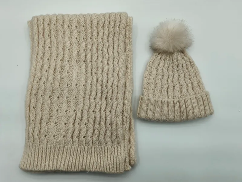 Cremefarbenes Winter-Set bestehend aus Zopfmuster-Beanie mit Kunstfell-Pompon und passendem Rippenstrickschal, flach ausgelegt