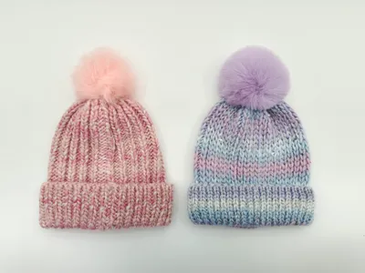 Zwei Grobstrick-Mützen mit Kunstfell-Pompons nebeneinander: eine in Rosa-Melange, die andere in Lavendel-Blau-Batik-Optik auf weißem Hintergrund