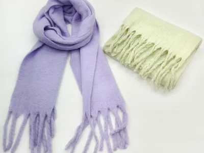 Zwei weiche Flauschschals mit Fransenenden: einer in Lavendel offen geloopt, einer in Mintgrün gefaltet, auf weißem Hintergrund
