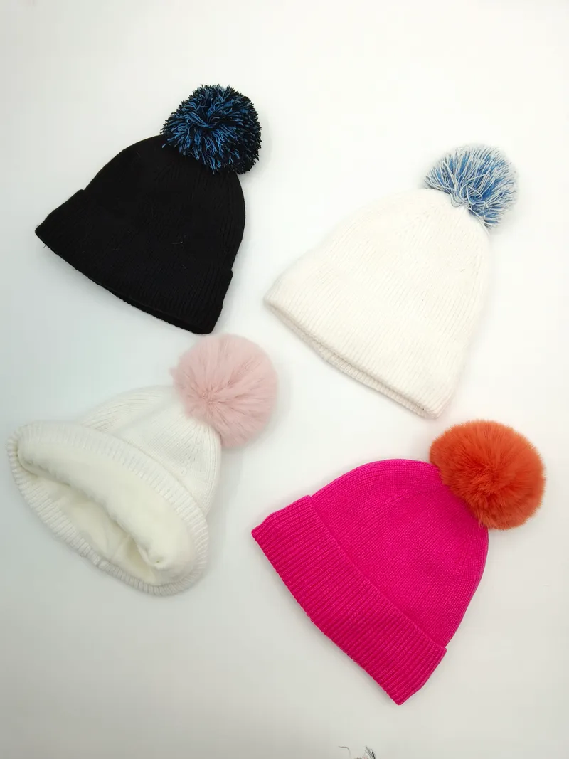 Vier Rippenstrickmützen mit bunten Bommeln in Schwarz, Weiß und Fuchsia-Pink, flach auf weißem Hintergrund angeordnet