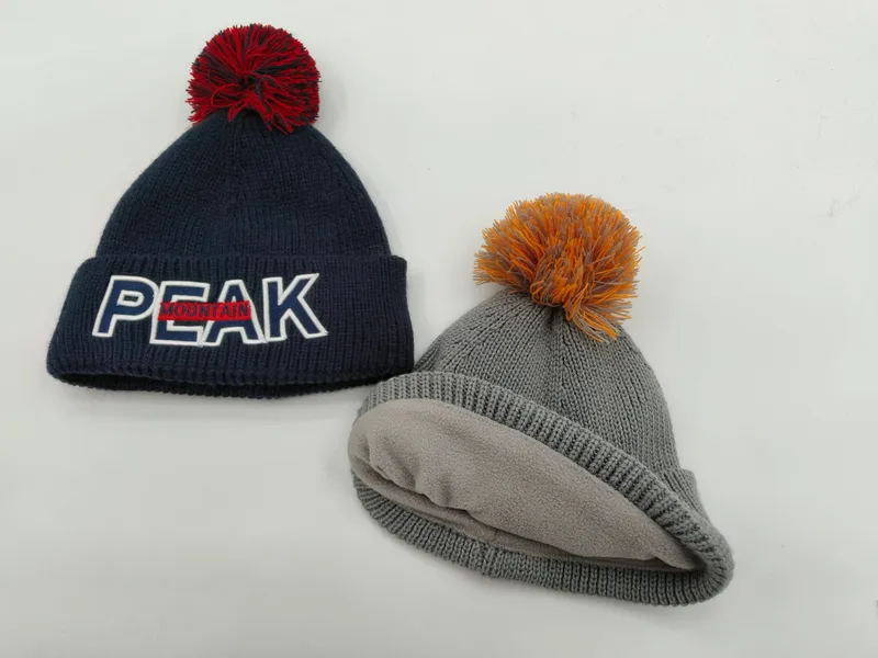 Zwei Strickmützen mit Bommeln: Navy-Blau mit Peak Mountain Logo und rotem Bommel, graue Fleece-gefütterte Mütze mit orangem Bommel, auf weißem Hintergrund