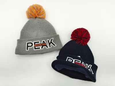 Zwei Grobstrick-Bommelmützen in Grau und Navy-Blau mit gesticktem Peak Mountain Abzeichen, auf weißem Hintergrund präsentiert