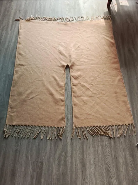 Flach liegender beiger Webponcho mit mittlerer Halsöffnung und gedrehten Fransen an den Rändern auf Holzboden
