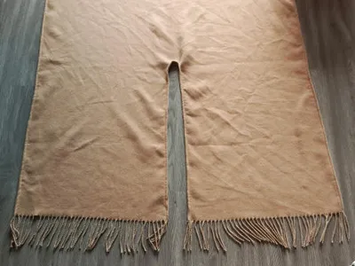 Flach liegender beiger Webponcho mit mittlerer Halsöffnung und gedrehten Fransen an den Rändern auf Holzboden