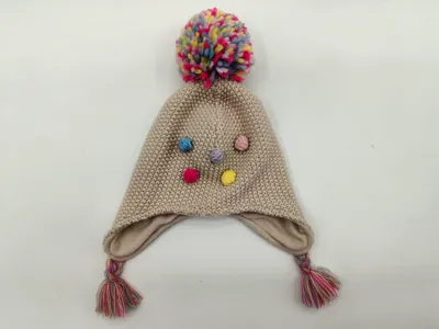 Beige gestrickte Kindermütze mit Ohrenklappen, großem buntem Bommel oben, farbigen Knopfakzenten und geflochtenen Zierquasten