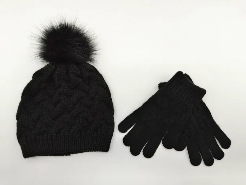 Schwarze Zopfstrick-Wintermütze mit fluffigem Kunstfell-Pompon und dazu passenden schwarzen Strickhandschuhen auf weißem Untergrund