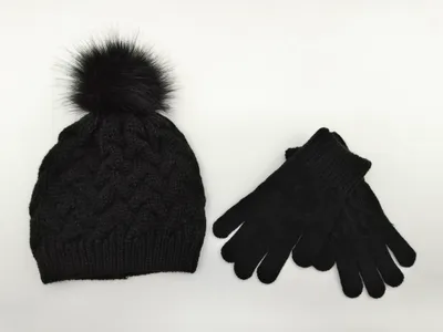 Schwarze Zopfstrick-Wintermütze mit fluffigem Kunstfell-Pompon und dazu passenden schwarzen Strickhandschuhen auf weißem Untergrund