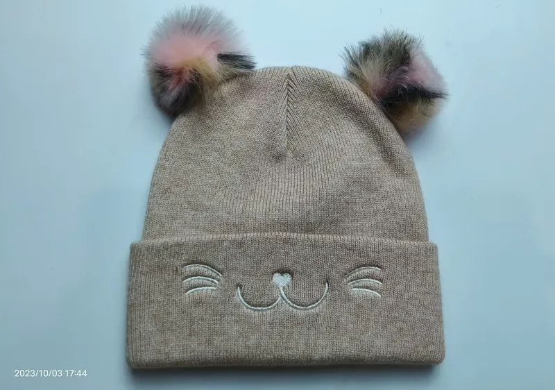Beigefarbene Kindermütze aus Strick mit zwei bunten Kunstfell-Pompons und aufgesticktem Katzengesicht auf der Umschlagkrempe