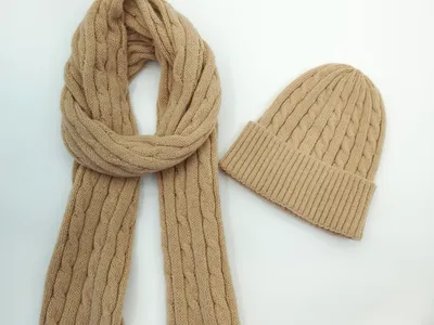 Kamelbrauner Zopfstrickschal aufgerollt neben passender Strickmütze auf weißem Untergrund