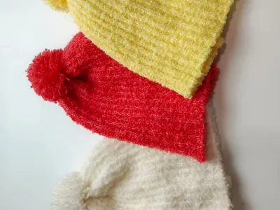 Drei flauschige Chenille-Strickmützen mit Bommel in Gelb, Rot und Weiß übereinandergestapelt vor weißem Hintergrund