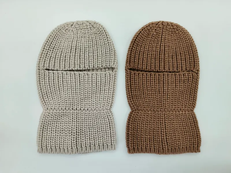 Zwei grob gestrickte Ganzkopfmasken flach liegend – eine in Hellbeige, eine in Warmbraun – mit Augenschlitz und Rippenstruktur