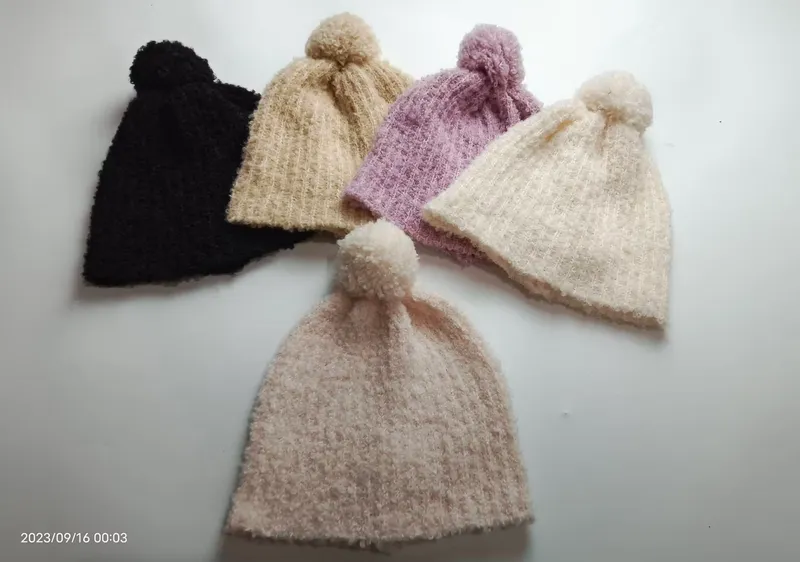Fünf weiche Wollmützen mit Bommeln in Schwarz, Beige, Flieder, Naturweiß und Rosé nebeneinander auf hellem Untergrund