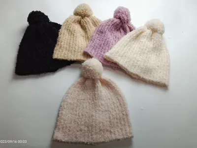 Fünf weiche Wollmützen mit Bommeln in Schwarz, Beige, Flieder, Naturweiß und Rosé nebeneinander auf hellem Untergrund