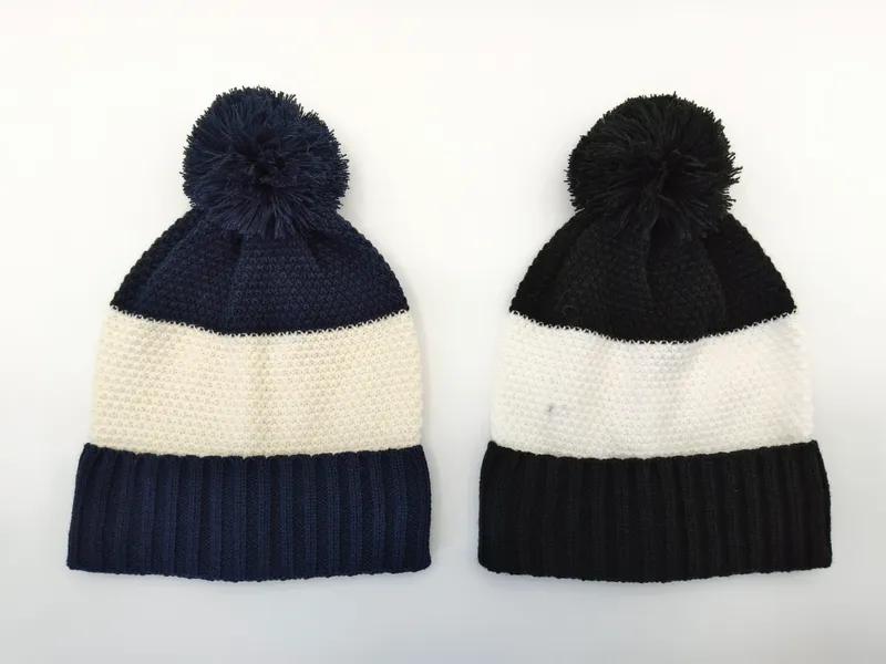 Zwei Rippenstrick-Pompom-Mützen nebeneinander – links Marine-Blau mit Cremstreifen, rechts Schwarz mit Cremstreifen – auf weißem Hintergrund