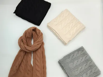 Vier gefaltete Strickschals mit Zopfmuster in den Farben Anthrazit, Elfenbein, Kamelbraun und Hellgrau auf hellem Untergrund, einer ausgebreitet in voller Länge