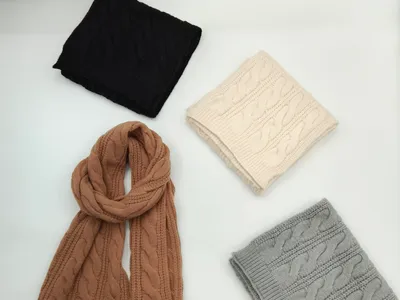 Vier Wollschals mit grobem Strickmuster in Schwarz, Cremeweiß, Sandbraun und Grau, sauber arrangiert auf weißem Hintergrund, einer vollständig entfaltet