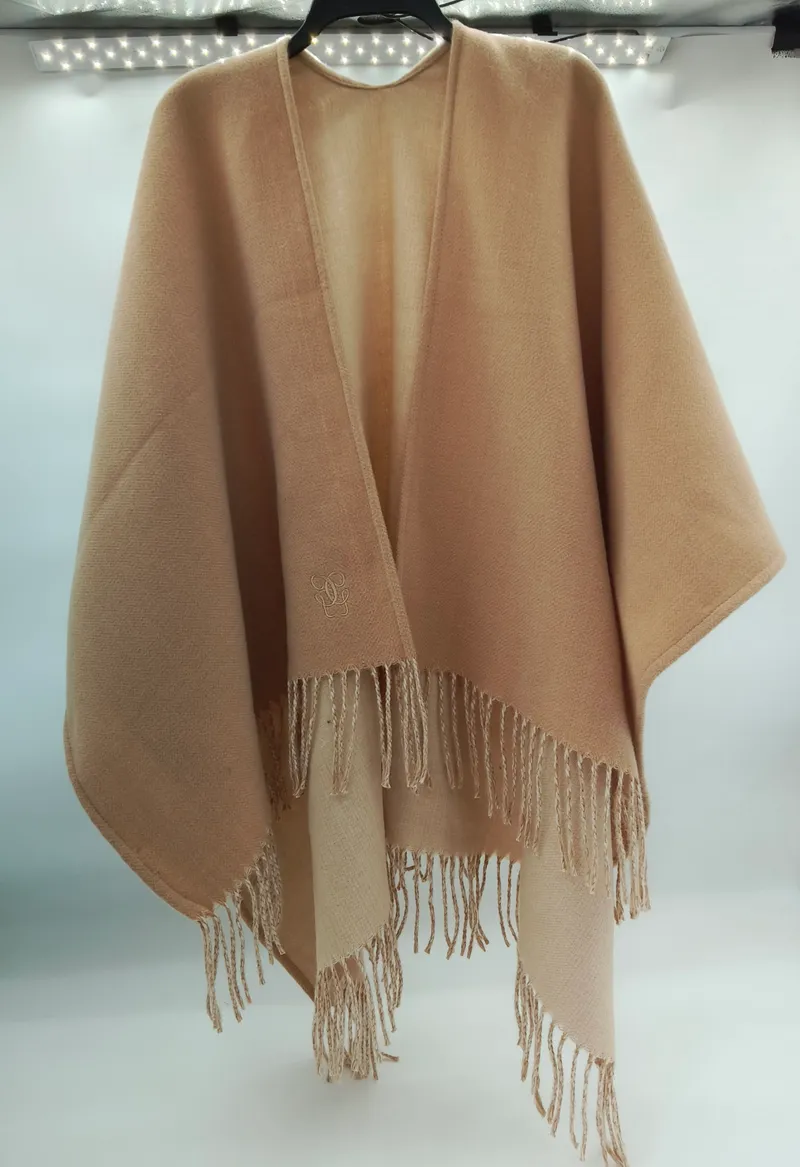 Sandfarbener Damenponcho mit Fransenbordüre und gesticktem Monogramm am Kragen, offen hängend auf Bügel
