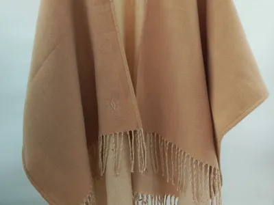 Sandfarbener Damenponcho mit Fransenbordüre und gesticktem Monogramm am Kragen, offen hängend auf Bügel