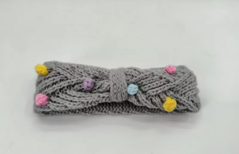 Gestricktes Stirnband in Grau mit Zopfmuster und farbenfrohen Mini-Pompons in Gelb, Rosa, Lila und Blau sowie dekorativem Schleifendetail in der Mitte