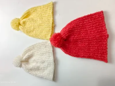 Drei gestrickte Bommelmützen in Sonnengelb, Korallenrot und Reinweiß nebeneinander liegend, flauschige Wintermützen im Flatlay