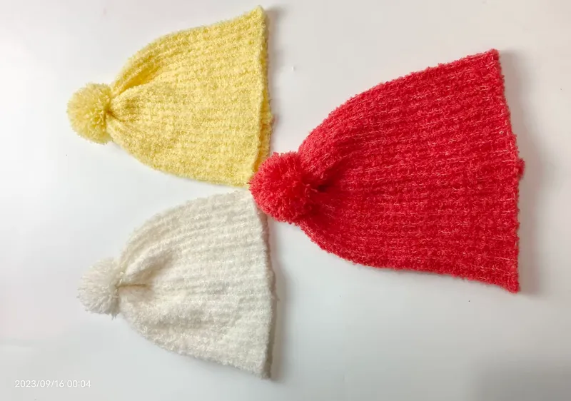Drei gestrickte Bommelmützen in Sonnengelb, Korallenrot und Reinweiß nebeneinander liegend, flauschige Wintermützen im Flatlay