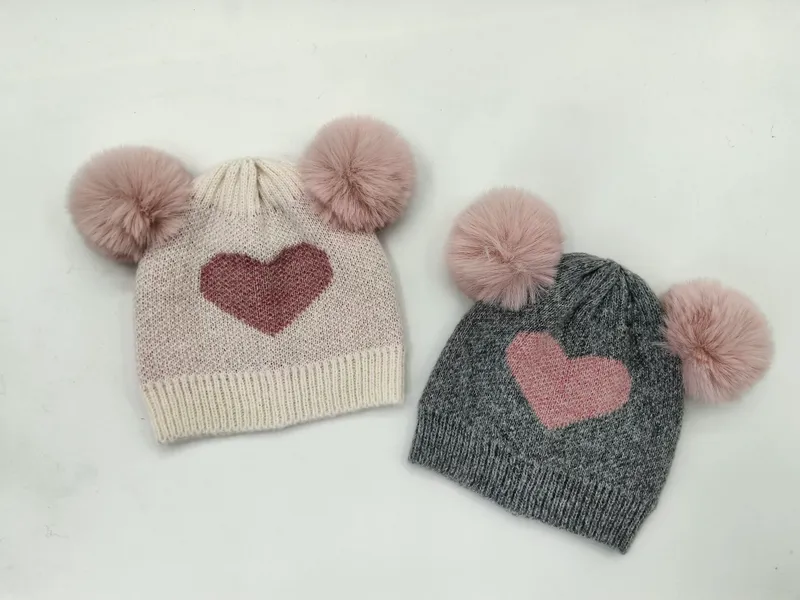 Flauschige Kindermützen aus Strickstoff in Creme und Grau mit zwei Kunstfell-Pompons und eingestricktem Herzmotiv, nebeneinander auf weißem Hintergrund
