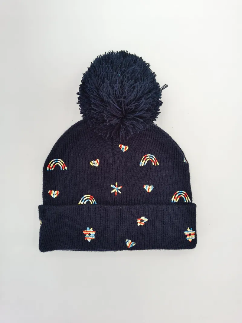 Marine-blaue gerippte Strickmütze mit großem Pompon und bunten gestickten Motiven wie Regenbogen, Herz und Stern