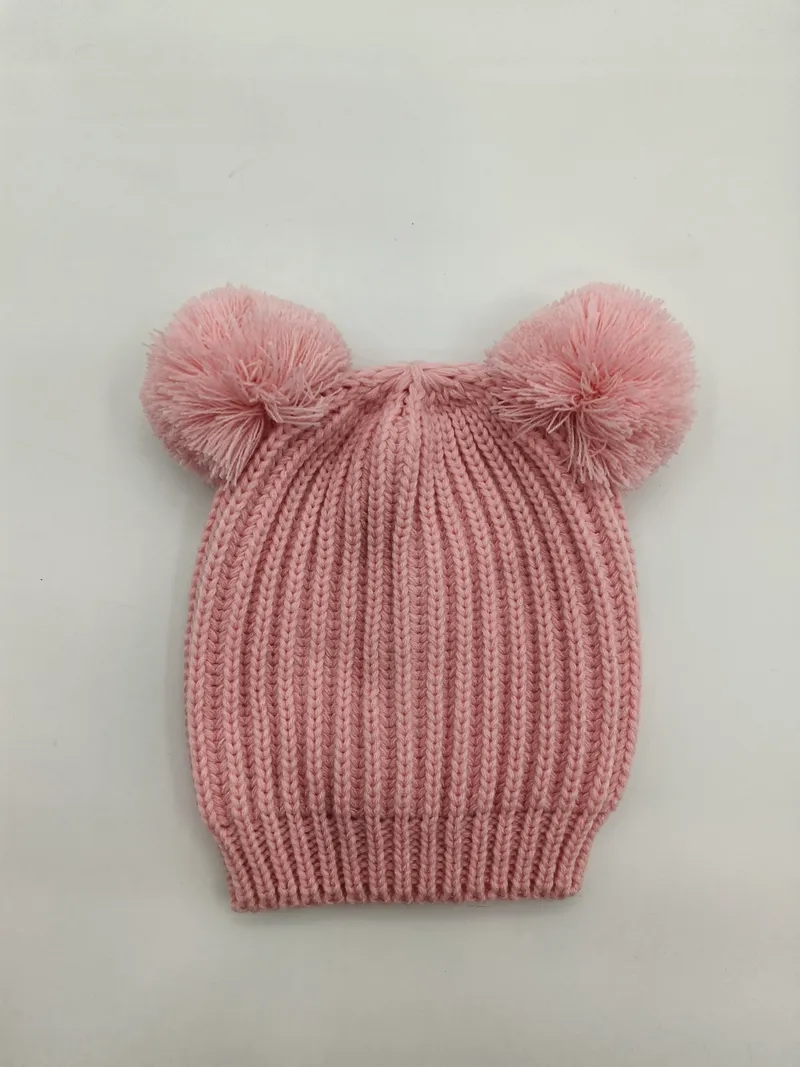 Rosa gerippte Babymütze mit zwei flauschigen Pompons oben, flach auf weißem Hintergrund liegend