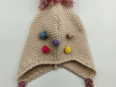 Handgestrickte Creme-Ohrenklappenmütze mit mehrfarbigem Pompon, kleinen bunten Garnbobbles und geflochtenen Quastenbändern mit Fransenden