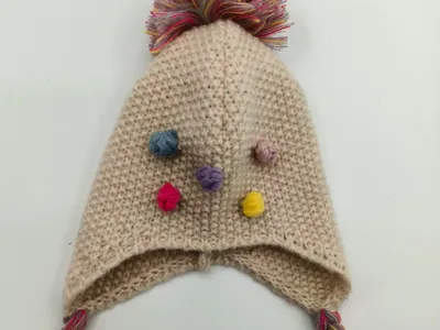 Handgestrickte Creme-Baby-Ohrenklappenmütze mit großem mehrfarbigem Pompon, kleinen bunten Mini-Pompons und geflochtenen Quastenbändern