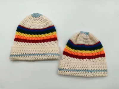 Zwei cremefarbene Häkelmützen mit bunten Regenbögen-Streifen in Blau, Grün, Gelb, Orange und Rot sowie hellblauem Bommel oben