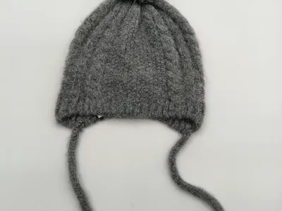 Hellgraue Babymütze aus grobem Zopfstrick mit rundem Wollbommel oben und zwei langen Kinnbändern aus Garn, freigestellt auf weißem Hintergrund