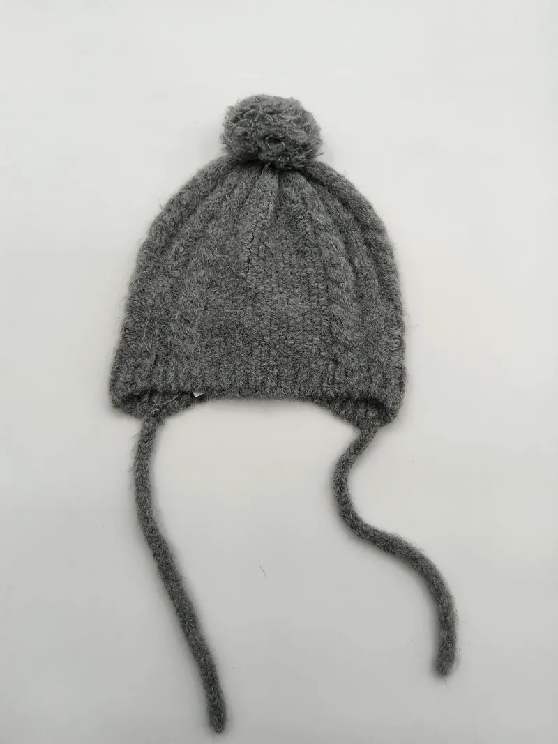 Hellgraue Babymütze aus grobem Zopfstrick mit rundem Wollbommel oben und zwei langen Kinnbändern aus Garn, freigestellt auf weißem Hintergrund