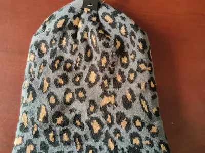 Grau-schwarze Strickmütze mit Leoparden-Tiermuster und beigen Flecken, auf einem Verkaufsbügel mit Etikett präsentiert