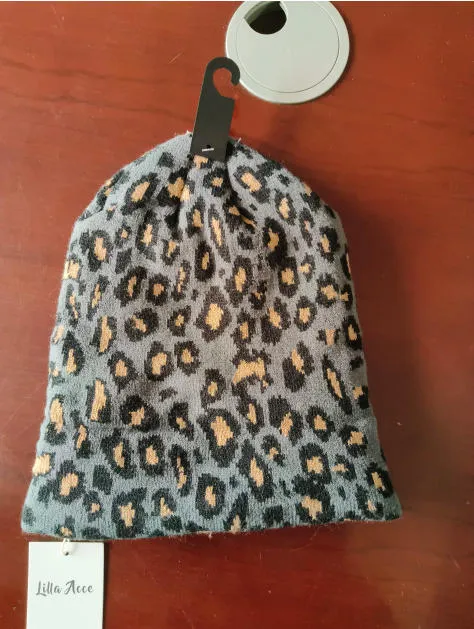 Grau-schwarze Strickmütze mit Leoparden-Tiermuster und beigen Flecken, auf einem Verkaufsbügel mit Etikett präsentiert