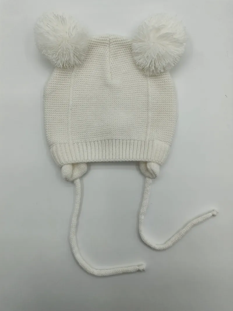 Weiße gestrickte Babymütze mit zwei flauschigen Bommeln oben und langen Kinnbändern, flach auf weißem Untergrund fotografiert