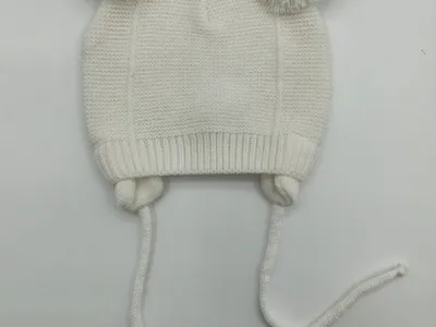 Weiße gestrickte Babymütze mit zwei flauschigen Bommeln oben und langen Kinnbändern, flach auf weißem Untergrund fotografiert