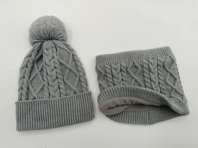 Graue Zopfstrick-Wintermütze mit Pompon und dazu passender Fleece-gefütterter Schlauchschal, freigestellt auf weißem Hintergrund