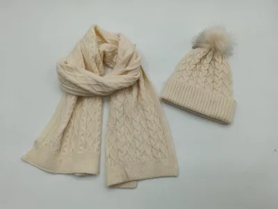 Cremefarbener Zopfstrick-Winterschal und passende Strickmütze mit Kunstfell-Pompon auf weißem Hintergrund