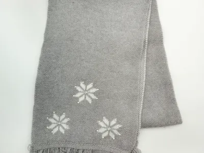 Gefaltete graue Strickdecke mit eingestrickten weißen Schneeflockenmotiven und Fransensaum auf weißem Untergrund