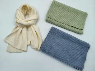 Drei winterliche Strickaccessoires in Creme, Salbeigrün und Stahlblau: locker gebundener langer Schal und zwei zusammengelegte Schlaufsnoods auf hellem Untergrund