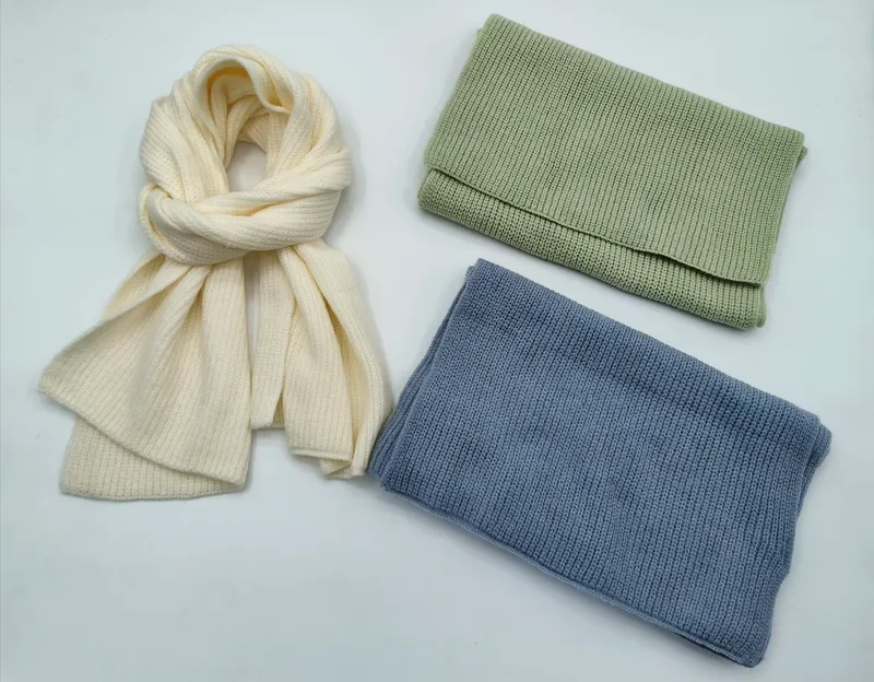 Drei winterliche Strickaccessoires in Creme, Salbeigrün und Stahlblau: locker gebundener langer Schal und zwei zusammengelegte Schlaufsnoods auf hellem Untergrund