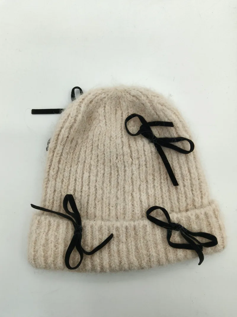 Dicke cremeweiße Wintermütze aus grobem Rippenstrick mit mehreren dekorativen schwarzen Samtbandschleifen, flach auf weißem Hintergrund fotografiert